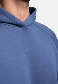 Nahaufnahme eines Halses und einer Schulter, die einen blauen Kapuzenpullover tragen. Der Stoff ist glatt und zeigt ein dezentes 2xu-Logo.