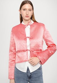 Vaatteiden yhdistelmä, jossa on kaunis light pink satiininen, vuorattu, lyhyt takki. Takissa on koristeelliset nauhat, pitkät hihat ja sileä pinta. Puvun alla on valkoinen paita.