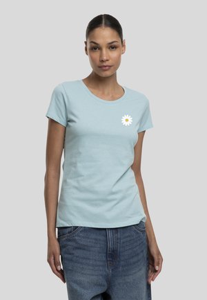 Femme aux cheveux foncés tirés en arrière porte un t-shirt bleu clair à manches courtes avec un petit motif marguerite blanche sur la poitrine et un jean bleu.