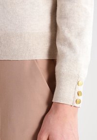 Béžový pletený svetr s žebrovanými lemy a třemi zlatými knoflíky jako ozdobou. Tkanina vypadá měkce s jemnou strukturou. Stylizováno s hnědými kalhotami.