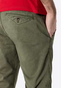 Olivgrüne Hose mit glatter Textur, ausgestattet mit einer Gesäßtasche mit dekorativer Naht und einem Metallelement. Eine Seitentasche ist sichtbar.