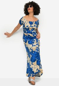 Niebieska i beżowa sukienka maxi w stylu tie-dye z odkrytymi ramionami, drapowanym gorsem i zwiewną sylwetką. Srebrne sandały dopełniają look.