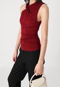 Burgundy sleeveless top with draped fabric and a twisted neckline, paired with black pants and a light beige handbag. Smooth texture.