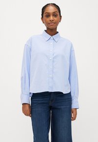 Šviesiai mėlynas, ilgarankovis marškinis su atšitiniu apykakle, turintis šiek tiek sutrumpintą dizainą ir saga užsegamus rankogalius, derinamas su tamsiomis džinsinėmis kelnėmis.