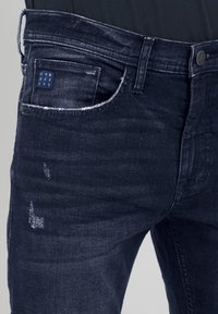 Denim jeans in dark blue, featuring a distressed texture with fading, a coin pocket, and a logo patch on the back pocket.