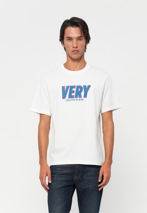 Man wearing white Calvin Klein t-shirt with blue and red "VERY" print, paired with dark blue jeans, standing against a plain white background.