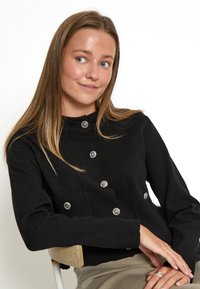 Melna adīta kardigāna ar augstu apkakli, sudraba pogu aizdari un divām priekšējām kabatām. Audums izskatās mīksts un teksturēts.