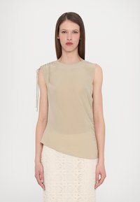 Top beige senza maniche con dettaglio arricciato sulla spalla sinistra, orlo asimmetrico e texture morbida e liscia. Abbinato a una gonna di pizzo.