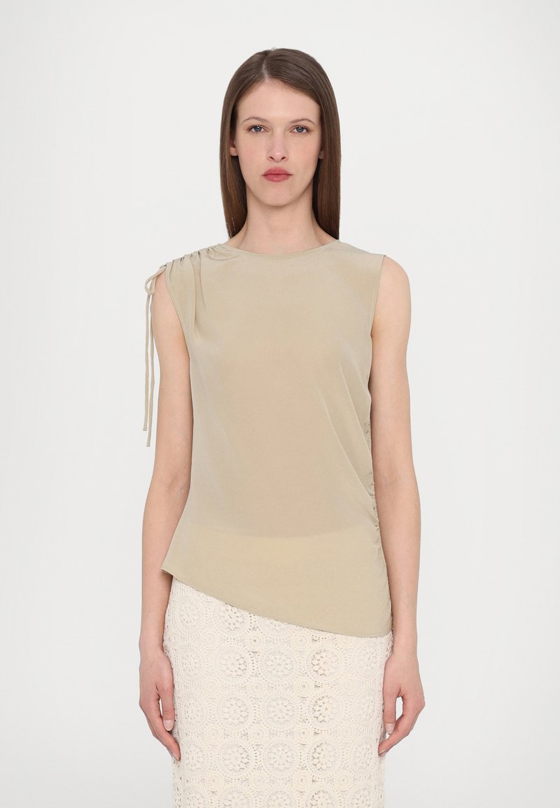 Top beige senza maniche con dettaglio arricciato sulla spalla sinistra, orlo asimmetrico e texture morbida e liscia. Abbinato a una gonna di pizzo.