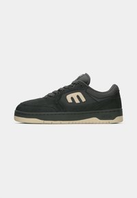 Black suede sneaker with rubber sole, padded collar, and lacing system. Features cream accents and a prominent logo on the side.