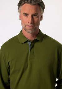 Grön pikétröja med en texturerad tyg, med en grå krage, tre knappar i placket och en logotyp på vänster bröst.