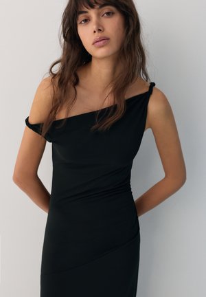 Fiatal nő hosszú barna hajjal, fekete, váll nélküli, testhezálló ruhát viselve, karjait a háta mögött tartva áll egy egyszerű háttér előtt.