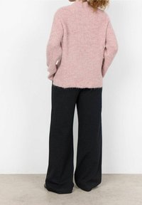 Pull en tricot rose clair à col montant, manches longues et texture côtelée, associé à un pantalon noir ample à motifs.