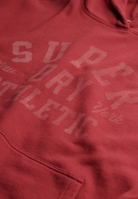 Czerwona bluza z kapturem z wyblakłym dużym nadrukiem tekstowym "Superdry Athletic" na przedniej części materiału.
