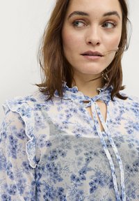 Leichte, durchsichtige Bluse in Blau mit floralem Muster. Verfügt über einen gerafften Kragen und eine Bindeschließe. Weiche, zarte Textur.