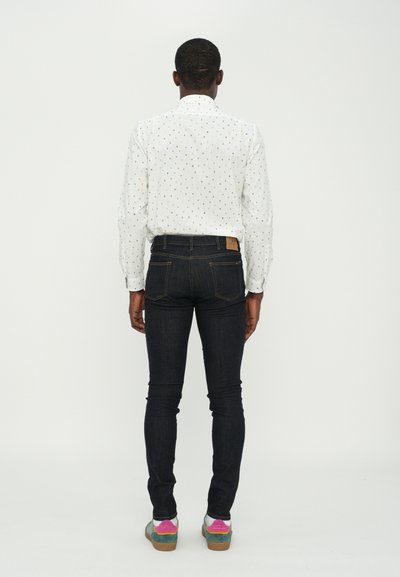 Chemise blanche avec de petits points multicolores, jeans bleu marine et baskets multicolores. Fond uni, couture visible sur le pantalon.