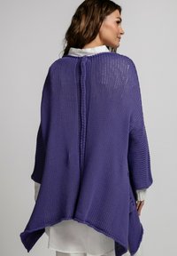 Fioletowy poncho dziane z luźnym, oversize'owym krojem, z teksturowanym wzorem i asymetrycznym dołem. Stylizowane na białej koszuli.