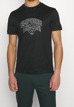 Mężczyzna noszący czarną koszulkę z krótkim rękawem z białym tekstem i logo "EMPORIO ARMANI" na piersi, zestawioną z ciemnymi spodniami.