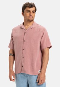 Koszula z krótkim rękawem, z kołnierzykiem w jasnoróżowym materiale, z teksturowanym wzorem i brązowymi guzikami. Logo metki widoczne na dolnym brzegu.