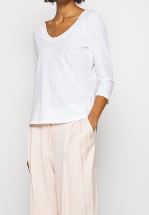 Femme portant un haut blanc à manches longues et col plongeant, ainsi qu'un pantalon beige clair à taille haute et jambes larges, une main dans la poche.