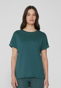 Tmavě zelené tričko s kulatým výstřihem, krátkými ohrnutými rukávy a mírně delším zadním lemem. Vyrobeno z hladké, měkké látky.