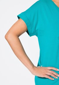 Top corto de color teal con un ajuste relajado, que muestra una textura suave y mangas amplias. La mano reposando en la cadera resalta la forma de la prenda.