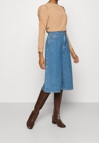Top beige a maniche lunghe con spalle arricciate abbinato a culotte in denim blu a gamba larga e stivali alti al ginocchio in pelle croccata marrone con tacco.