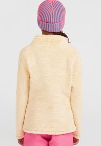 Cremefarbene Fleecejacke mit einer weichen, strukturierten Oberfläche und hohem Kragen, von hinten betrachtet, kombiniert mit einer gestreiften Beanie in Rosa und Blau.