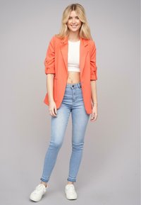 Koronkowa marynarka na białym topie crop, w połączeniu z jasnoniebieskimi slim fit dżinsami i białymi sneakersami. Marynarka ma podwinięte rękawy i dopasowany krój.