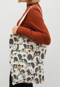 En person holder en tote bag med en mængde tegneseriepingviner iført hatte og tørklæder, mod en lysegrå baggrund.