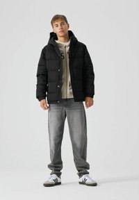 Giacca nera imbottita con cappuccio, felpa beige sotto, jeans grigi e sneakers bianche con dettagli neri. Design semplice e funzionale.