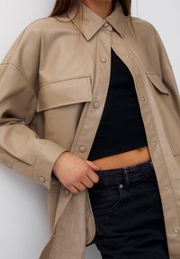 Veste-chemise en similicuir beige à coupe décontractée, boutons-pression, et poche poitrine, associée à un haut côtelé noir et un jean foncé.