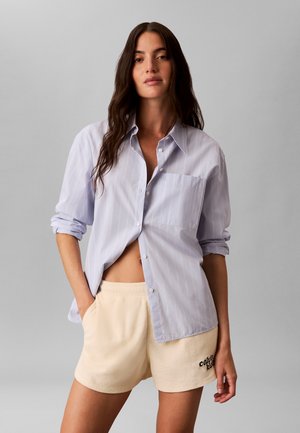 Junge Frau mit langen dunklen Haaren trägt ein hellblaues Hemd mit Knöpfen und beige Shorts mit dem Schriftzug „Calvin Klein“ auf dem linken Bein, steht mit den Händen in den Taschen.