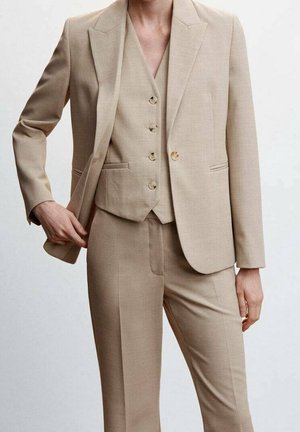 Beżowy garnitur szyty na miarę noszony z dopasowaną zapinaną kamizelką i spodniami, model koryguje marynarkę lewą ręką, jednolite jasne tło.