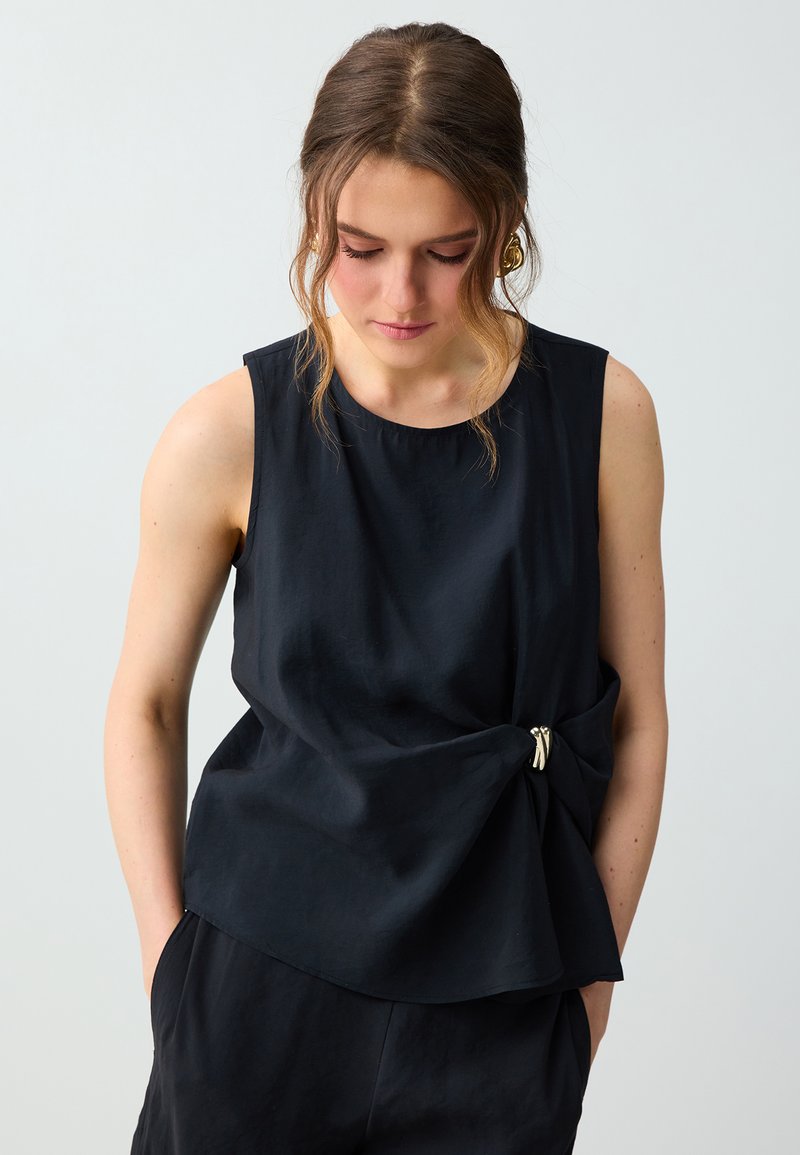 Femme aux cheveux ondulés portant un haut noir sans manches avec un nœud sur le côté et une broche dorée, regardant vers le bas, les mains dans les poches.
