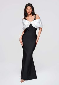 Robe de soirée noire à la silhouette ajustée, bretelles spaghetti, et un cache-cœur en satin blanc avec une boucle décorative. Sandales à talons transparents.