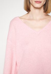 Femme portant un pull rose doux à col en V avec un motif en maille lâche, montrant le décolleté et une partie des cheveux châtain clair.