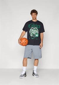 Černé tričko se zeleným potiskem a logem "Celtics", doplněné šedými basketbalovými šortkami a bílými ponožkami. Drží oranžový basketbalový míč.