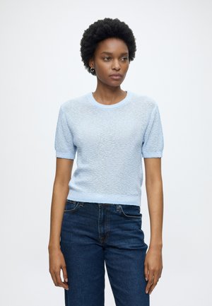 Femme aux cheveux courts et bouclés portant un pull léger bleu clair texturé à manches courtes et un jean bleu foncé, debout devant un fond blanc uni.