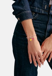 Silberkette mit gelbem Smiley- und pinkfarbenem Herzanhänger, detailliert in Emaille, getragen an einer Hand, die auf Denimstoff ruht.