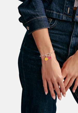 Silberkette mit gelbem Smiley- und pinkfarbenem Herzanhänger, detailliert in Emaille, getragen an einer Hand, die auf Denimstoff ruht.