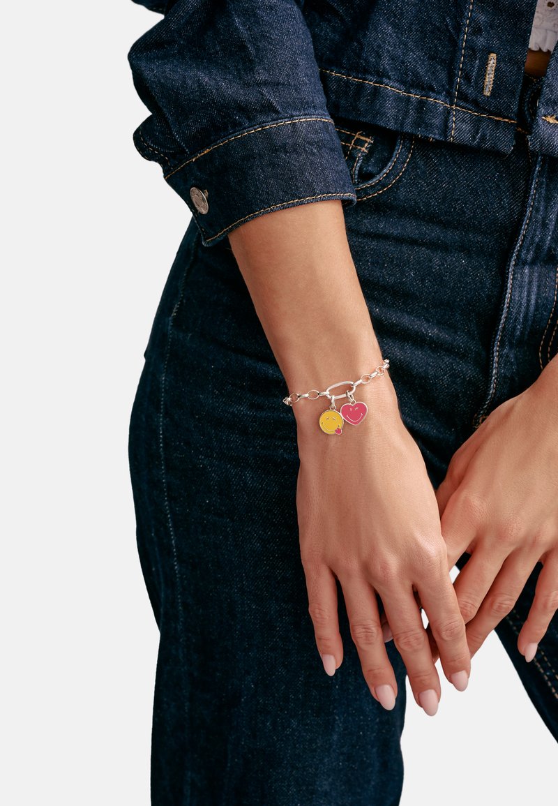 Silberkette mit gelbem Smiley- und pinkfarbenem Herzanhänger, detailliert in Emaille, getragen an einer Hand, die auf Denimstoff ruht.