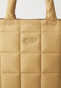 Quilted, tan tote bag with a soft texture. Rounded handles and a subtle logo in relief. Stitch detailing adds dimension to the design.