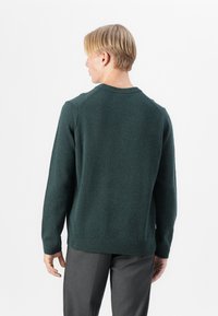 Pulover tricotat de culoare verde închis, cu guler rotund, manșete și tiv cu nervuri. Prezintă o textură netedă și un design ajustat. Purtați-l împreună cu pantaloni gri.