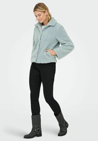 Light blue, textured fleece jacket with a collar, front button closure, and side pockets. Worn with black pants and gray boots.