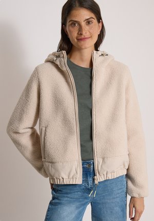 Femme aux cheveux foncés portant une veste polaire beige zippée par-dessus une chemise verte et un jean bleu, debout avec une main dans la poche.