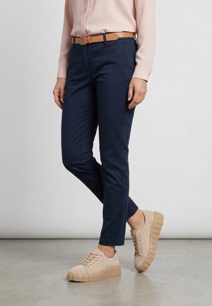 Persoon draagt marineblauwe slim-fit broek, bruine riem, lichtroze blouse en beige platformsneakers, staand met één been licht gebogen.
