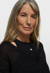 Zwarte gebreide trui met een losse pasvorm en een V-vormige halslijn, gedragen over een zwart topje met een rechthoekige logopatch. Lang, golvend grijs haar.