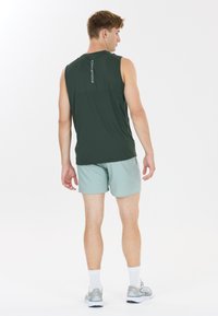Homme portant un haut sans manches vert foncé avec le texte « ENDURANCE », un short vert clair, des chaussettes blanches et des chaussures de course grises, debout de dos.
