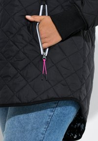 Giacca imbottita nera con orlo asimmetrico, dotata di tasca con zip e coulisse rosa e polsini a coste. Abbinata a jeans blu.